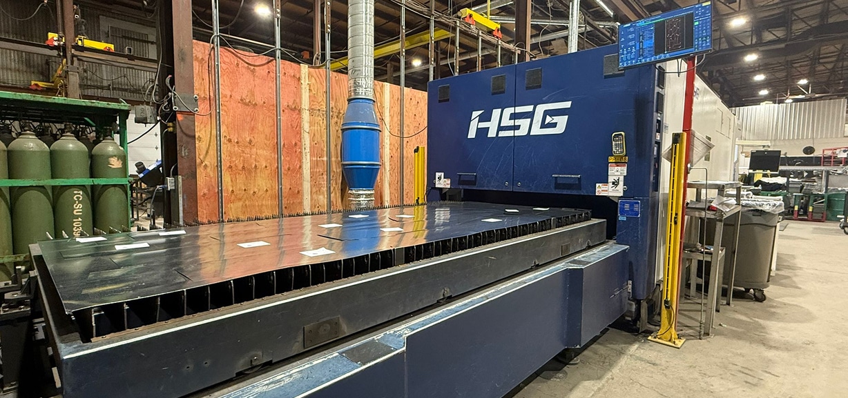 Une grande machine industrielle bleue de marque HSG dans une usine, avec des tôles métalliques sur sa plate-forme. On aperçoit à l'arrière-plan des bouteilles de gaz et divers équipements, ainsi qu'une cloison en bois et de hauts plafonds.