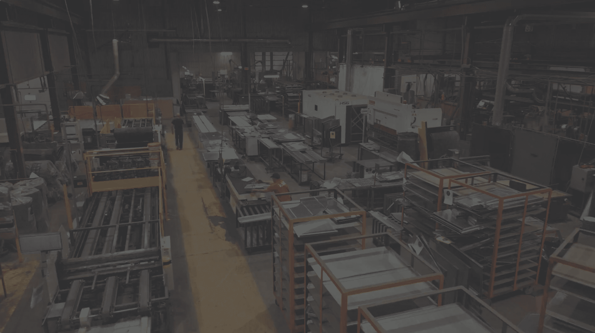 Intérieur d'une grande usine industrielle, avec des rayonnages métalliques, des machines, des établis et du matériel éparpillé un peu partout. L'espace est faiblement éclairé ; on aperçoit quelques ouvriers parmi les machines et les postes de travail.