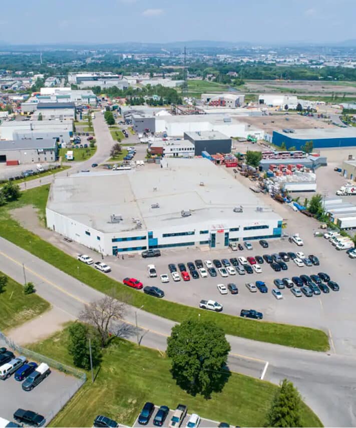 Vue aérienne d'une zone industrielle comprenant de grands entrepôts blancs, un parking rempli de voitures, des espaces verts, des routes et, au loin, un paysage urbain sous un ciel partiellement nuageux.