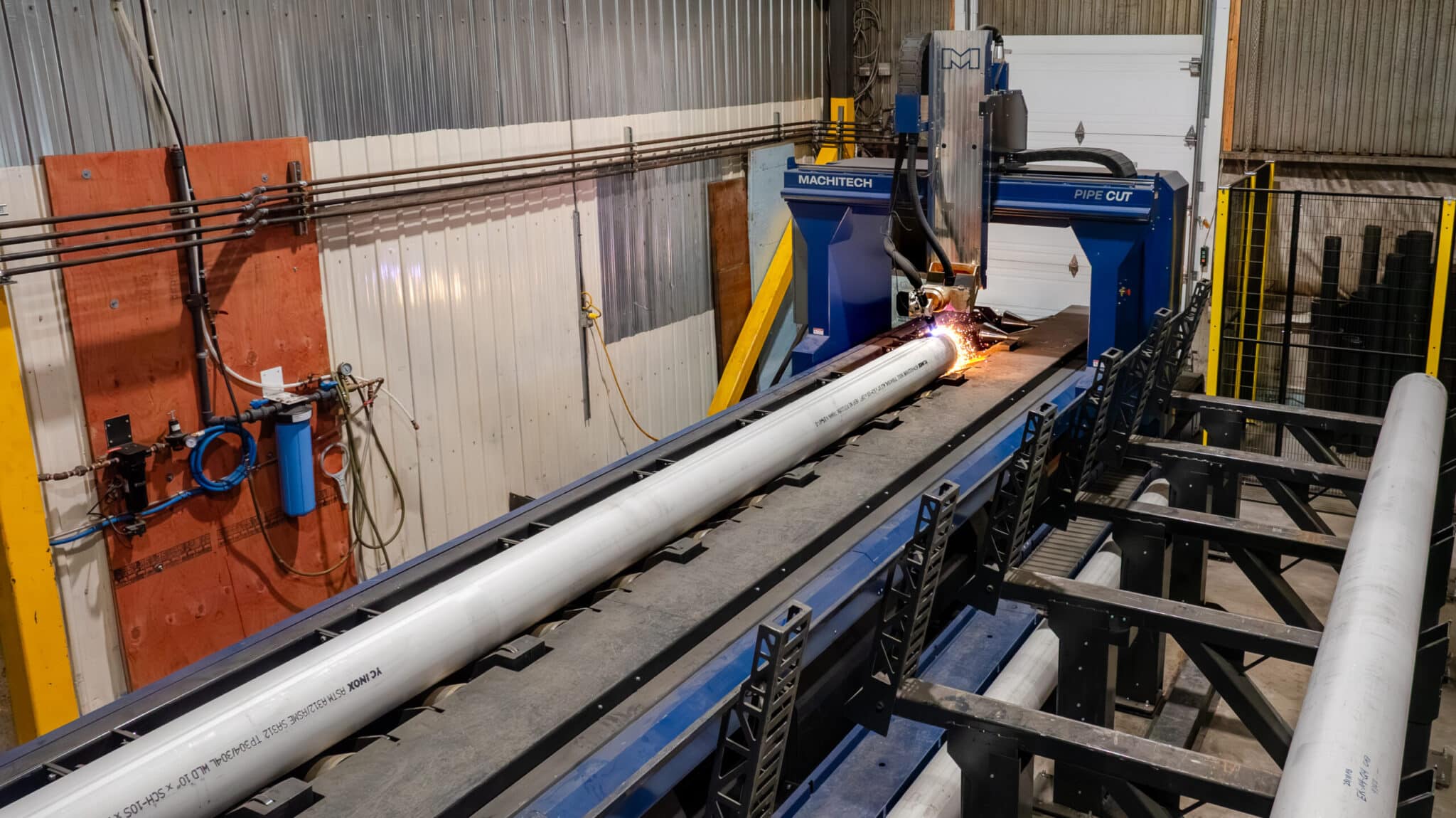 Une machine à commande numérique découpe un long tuyau métallique, projetant des étincelles brillantes, dans un décor d'usine industrielle où l'on aperçoit des murs métalliques et des équipements.