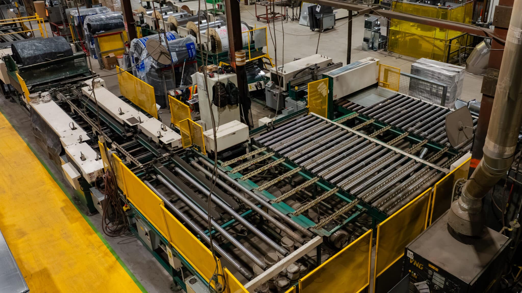 Un atelier équipé de grosses machines industrielles et de convoyeurs à rouleaux, entouré de barrières de sécurité jaunes, où sont traités des matériaux dans un vaste espace de production.