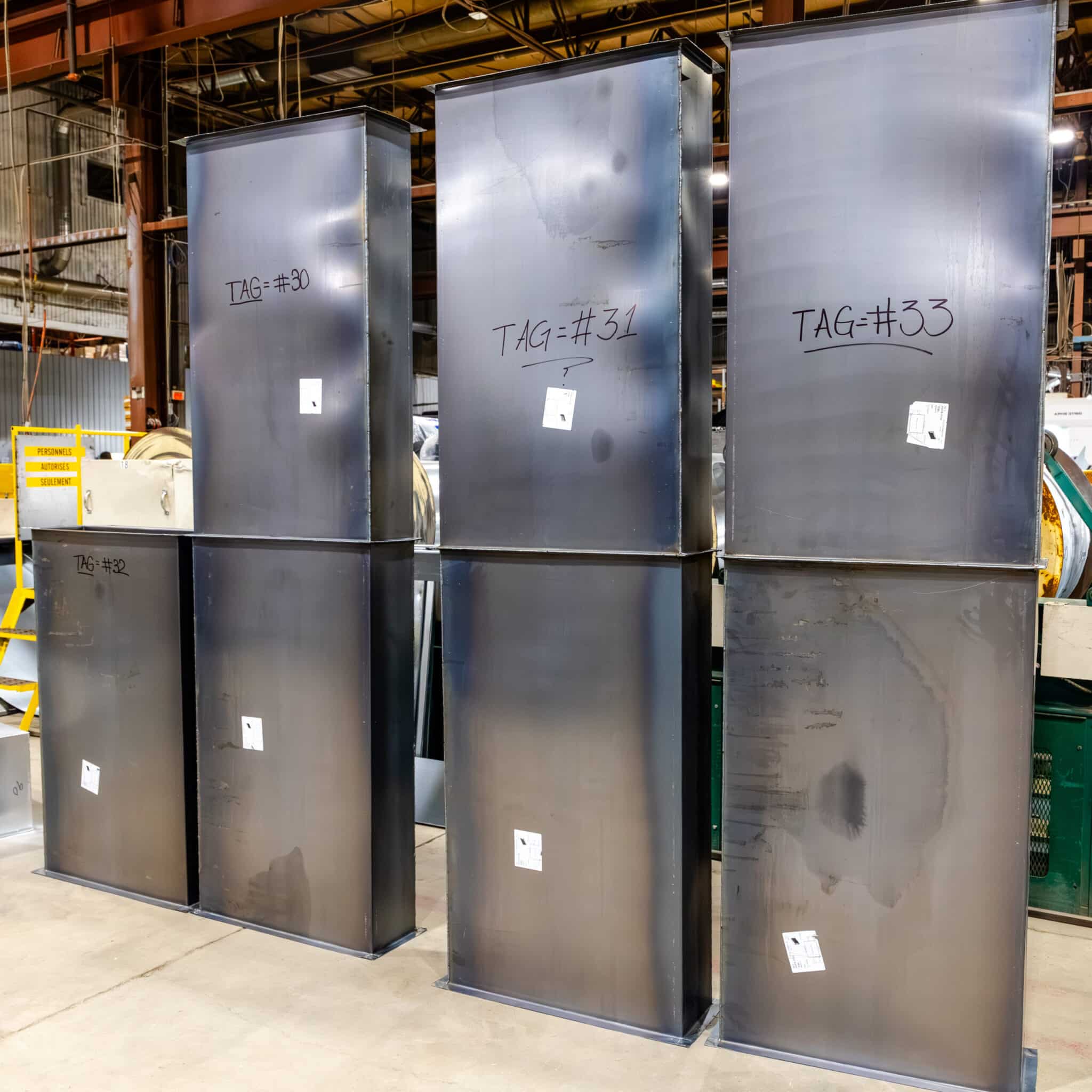 Quatre grands coffrets électriques métalliques sont alignés côte à côte dans un entrepôt industriel ; chacun porte une étiquette sur laquelle figure un numéro inscrit au marqueur noir. À l'arrière-plan, on aperçoit du matériel, des garde-corps jaunes et une affiche présentant une offre de services.