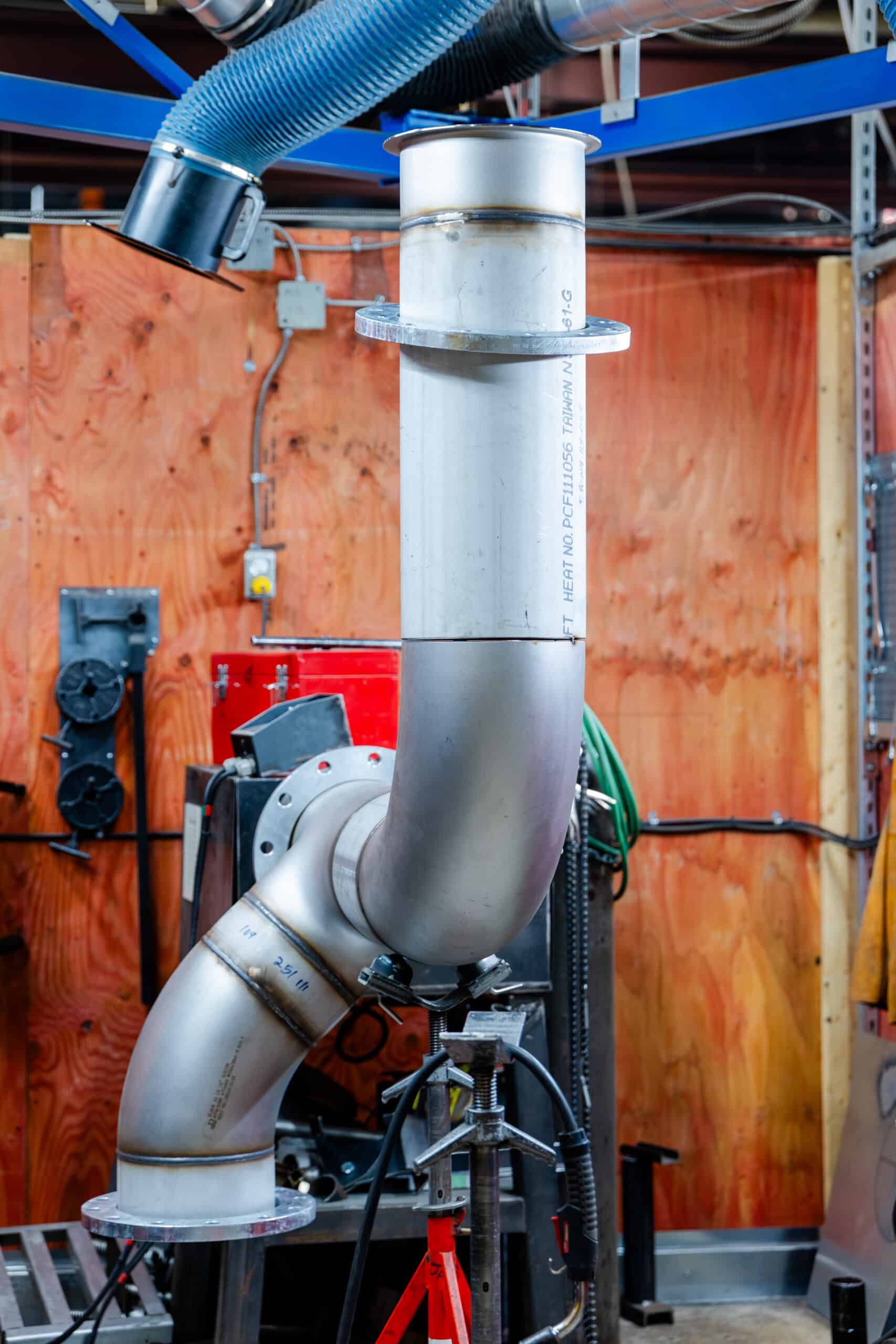 Un grand ensemble de tuyaux métalliques comportant plusieurs coudes est monté verticalement sur un support dans un atelier industriel, illustrant la gamme de services proposés, avec des murs lambrissés, des coffrets électriques et un grand tuyau de ventilation bleu suspendu au plafond.
