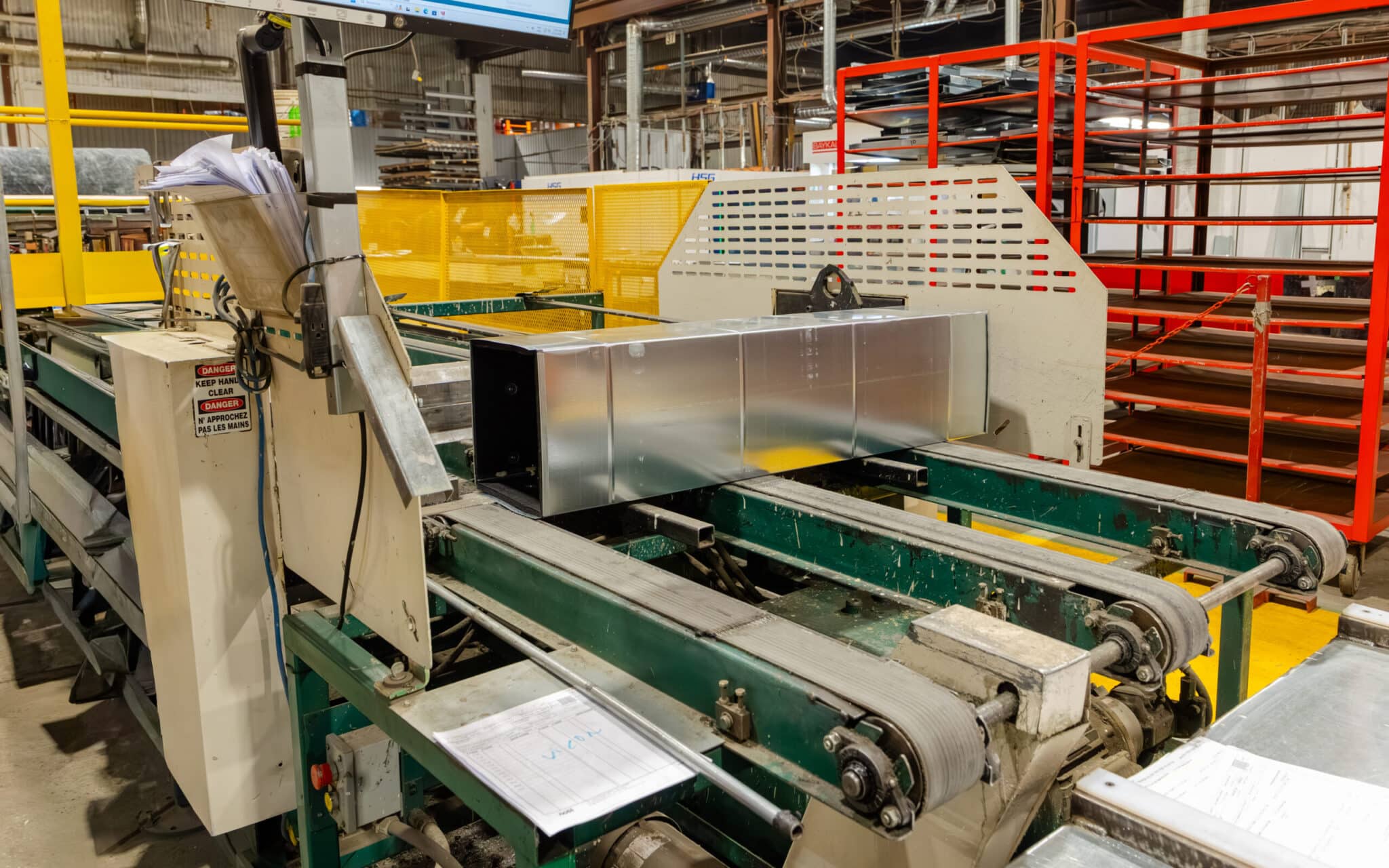 Un conduit métallique est en cours de fabrication sur une chaîne de montage automatisée dans une usine, au milieu de machines, de tapis roulants et de piles de documents papier.
