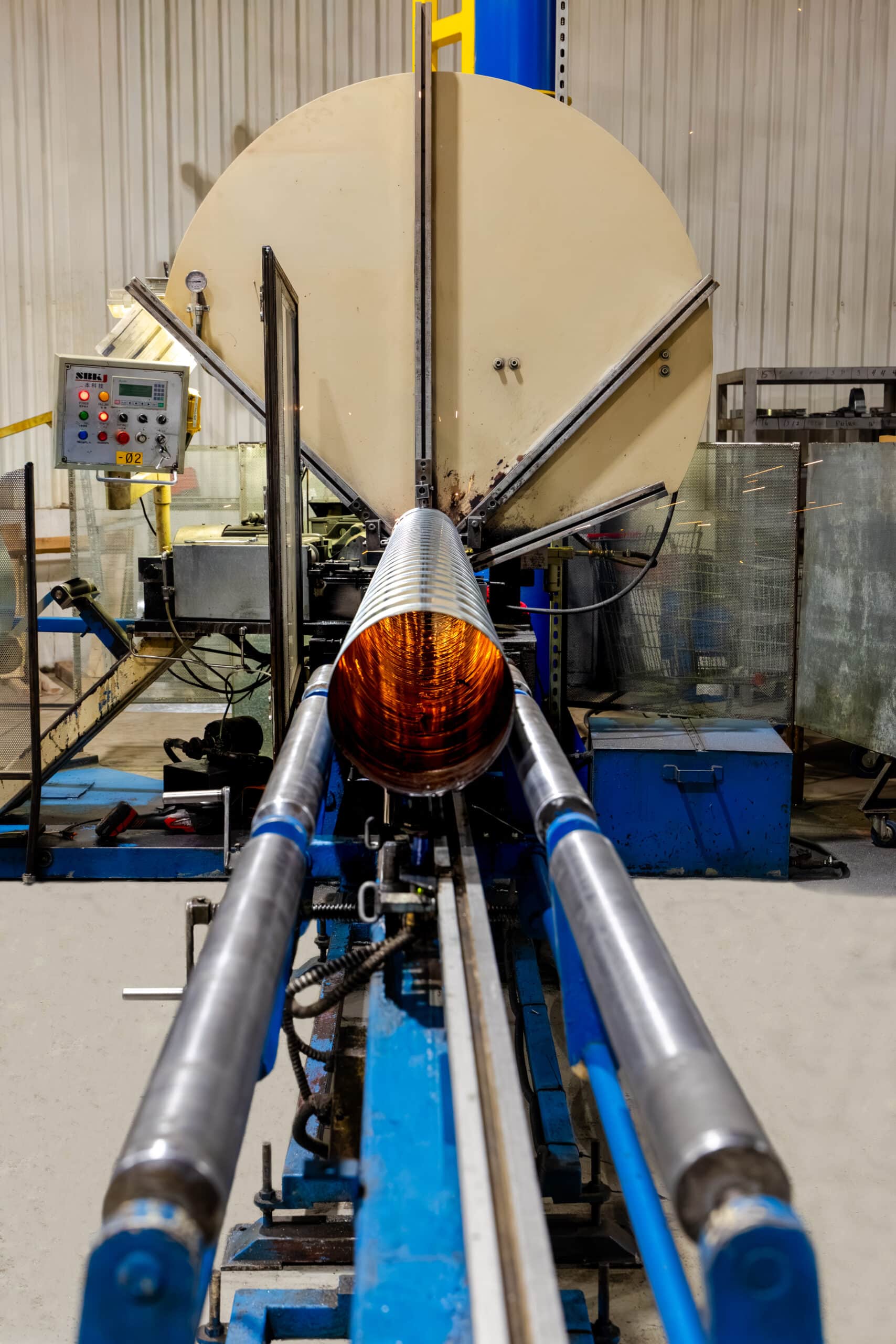 Dans un atelier, une grande machine industrielle enroule du fil métallique autour d'un mandrin cylindrique en rotation ; on aperçoit sur le côté des tiges métalliques qui guident le matériau ainsi que des panneaux de commande.