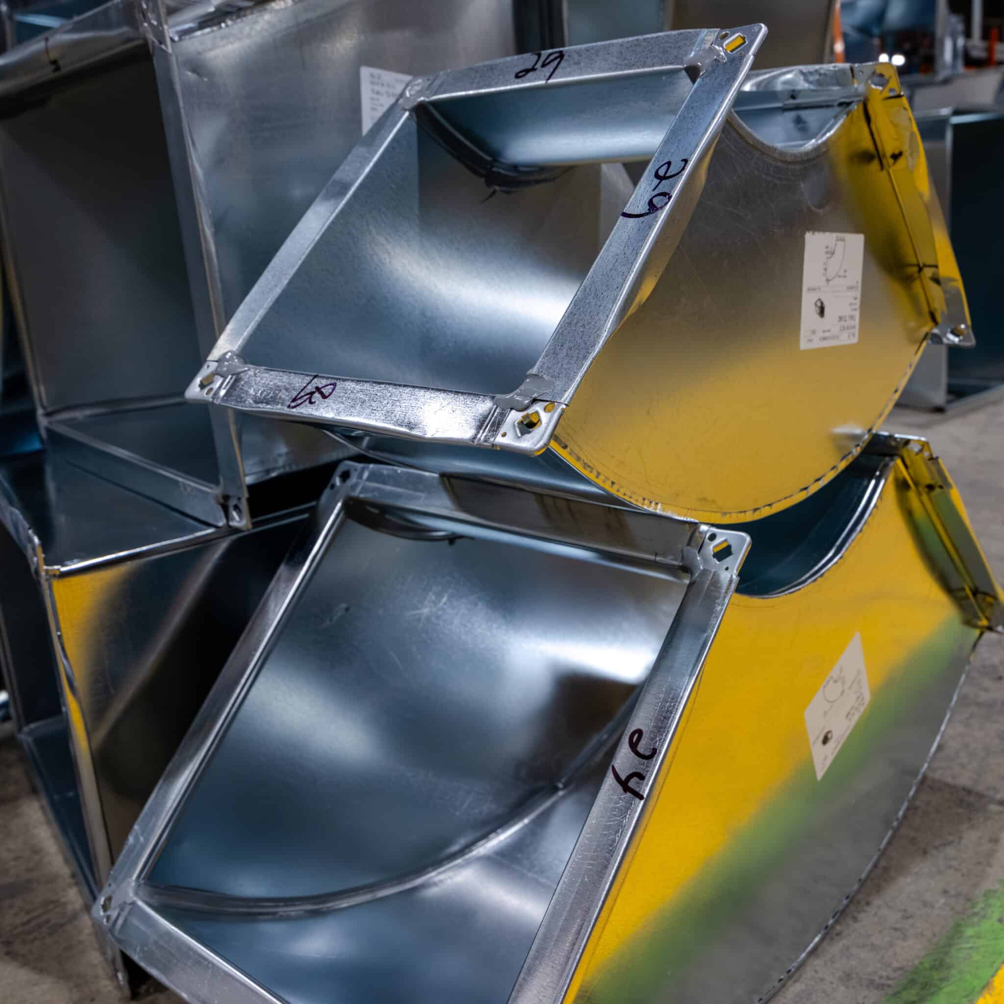 Des sections de conduits de climatisation en métal brillant empilées, portant des annotations manuscrites aux angles, disposées sur un sol en béton dans un environnement industriel, prêtes à être intégrées dans le cadre d'une offre de services.