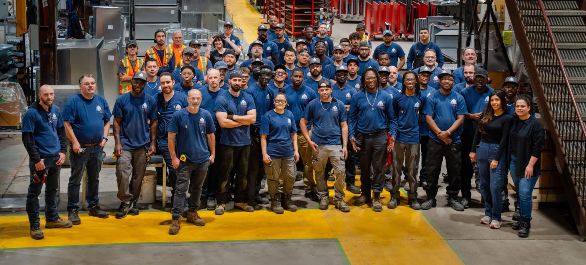 Un groupe important d'employés d'entrepôt, vêtus de chemises bleues identiques, posent ensemble à l'intérieur d'un vaste bâtiment industriel, debout sur un sol marqué de bandes jaunes, près d'équipements métalliques et d'escaliers.