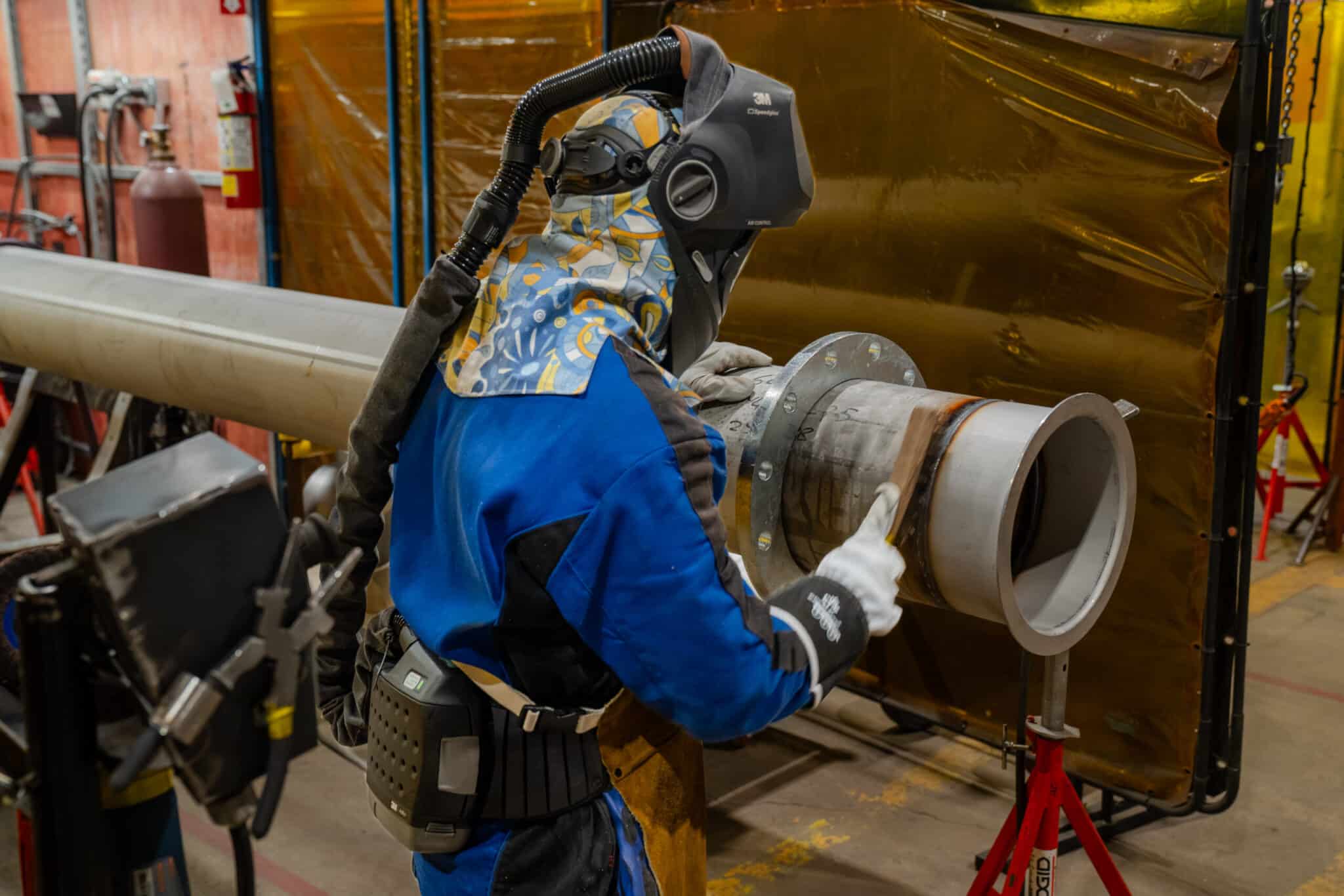 Un ouvrier, équipé d'un équipement de protection et d'un masque respiratoire, inspecte un grand tuyau métallique dans un atelier industriel, entouré de matériel métallique et d'un écran de sécurité jaune, illustrant ainsi l'offre de services de l'entreprise en matière de sécurité et d'inspection industrielles.