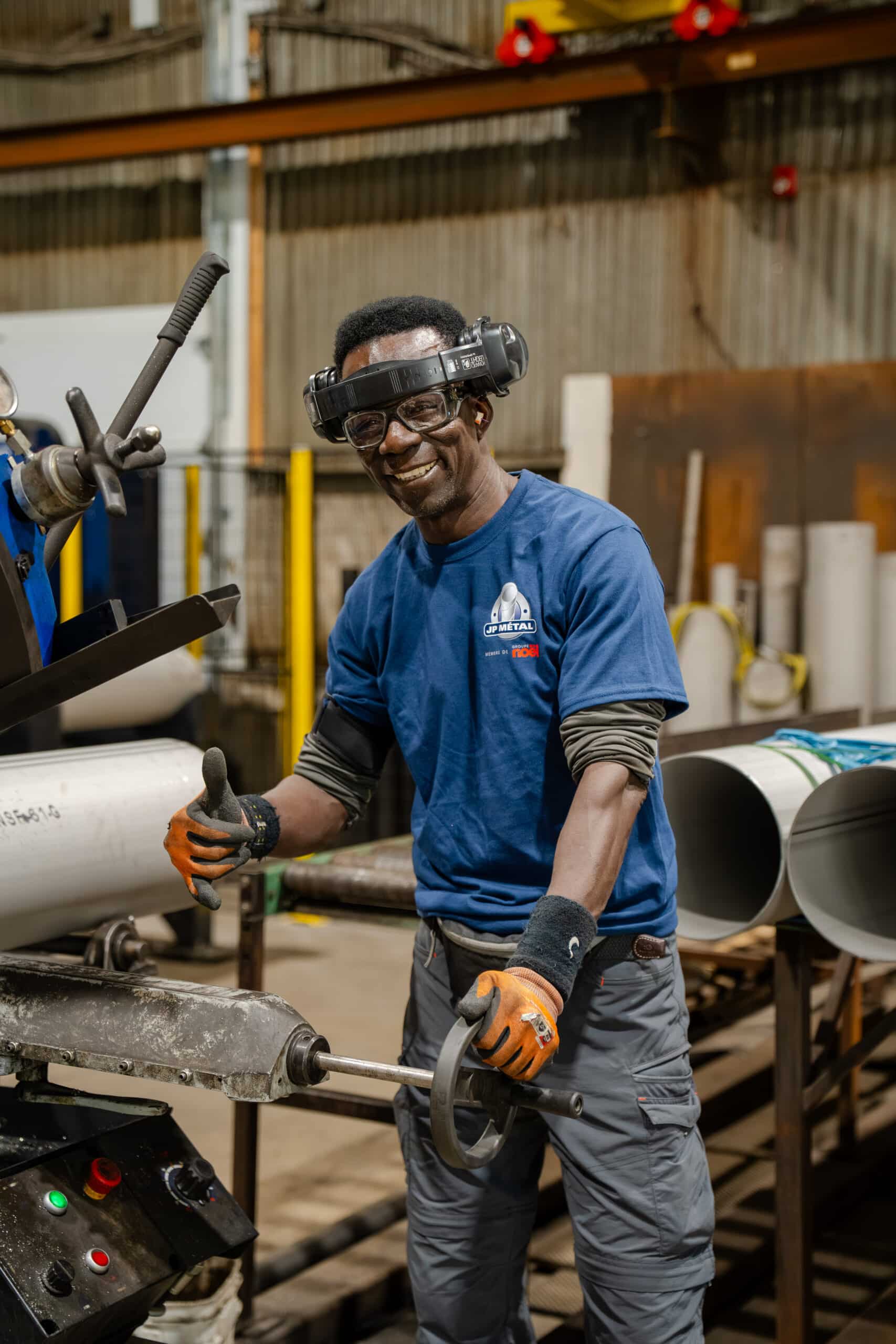 Un ouvrier souriant, portant des lunettes de protection, des gants et une chemise bleue, lève le pouce tout en manipulant un équipement industriel dans une usine, avec en arrière-plan de gros tuyaux et des structures métalliques.